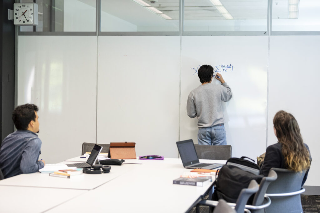 Photo of a person writing on a whiteboard while two others look on in a collaborative learning or a study group session