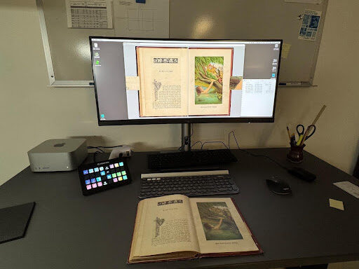 Image is of a computer monitor with a colorful keypad and keyboard on a black desk with a book opened to an illustrated page in the foreground. On the monitor the same page spread image is opened to show how quality control is performed for items that have been digitized. The book is titled Märchen Garten and is a children's fairytale book published in 1880. The text is German fraktur style with stylized chapter headings and very embellished drop caps. The image on the right page is a color depiction of a scene from the story Puss in Boots. The digitizer will view the digital images and compare them to the physical book to ensure accuracy.