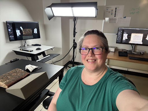 Image of a female figure (Me, Michelle) taking a selfie in the foreground with a physical book sitting on a black table on the left. Out of frame above the book is a digitization camera pointing down to take pictures of the book. In the left background a monitor shows the live image from the camera with the closed book, page weights, and support foam. In the right background, another monitor shows the last photo taken of the book that has not been rotated or cropped for public view. Right behind the figure (me) is a rectangle camera light and out of frame on the other side of the table is an identical light so the objects can be lighted without shadows