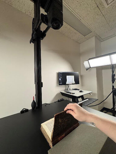 Image is of a camera in the upper foreground pointing down on a book on a black table held open by the hand of the picture-taker and resting against a piece of foam support. In the background a computer monitor on a little table which shows the live camera image of the book. To the right of the monitor is a wide rectangle studio light. The book is a 14th-century Latin manuscript written on vellum and in the process of digitization for wider user access.
